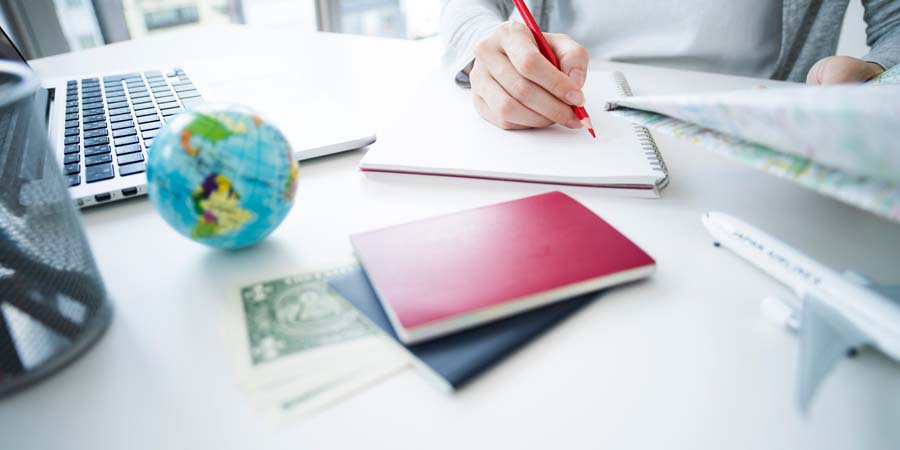 留学の計画を立てる人のデスク。ノートにメモを取る手元、地球儀、パスポート、1ドル札、ノートパソコン、飛行機の模型が机上に置かれている