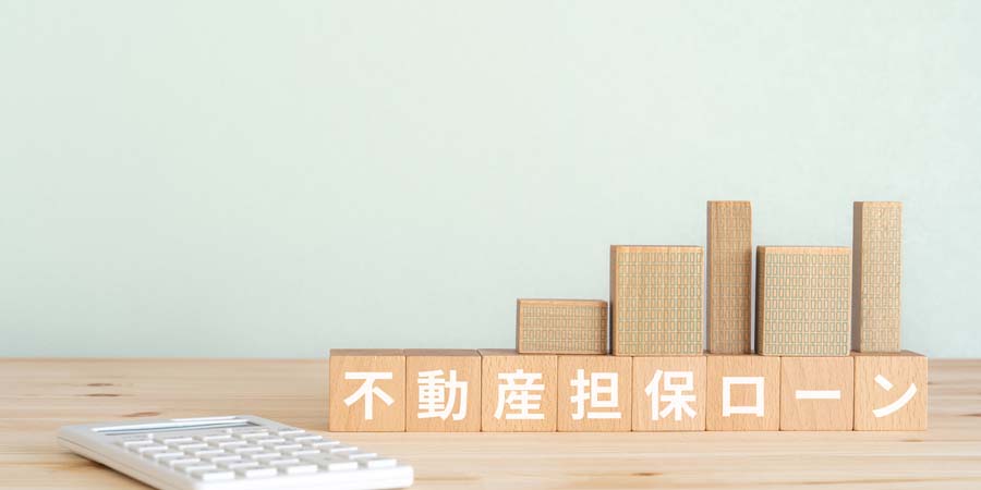木製ブロックで構成されたビルの模型と、手前に『不動産担保ローン』の文字が書かれたブロックが並ぶ