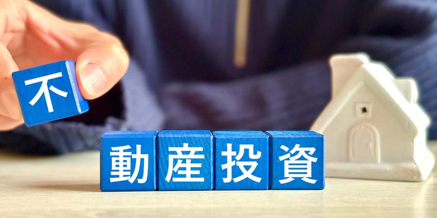 『不動産投資』の文字が書かれた青いブロックを並べる手元と、背景には白い家の模型が置かれている