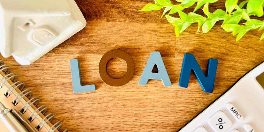 木製のテーブルの上に『LOAN』の文字が並べられ、周囲に家の模型、ノート、電卓、観葉植物が置かれている