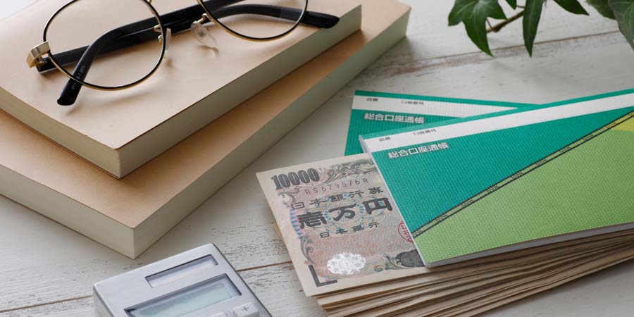 通帳と1万円札、電卓、メガネ、ノートが並ぶ写真。教育費の管理や進学資金の準備をイメージ