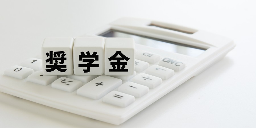 「奨学金」と書かれた3つのサイコロが電卓の上に置かれている画像